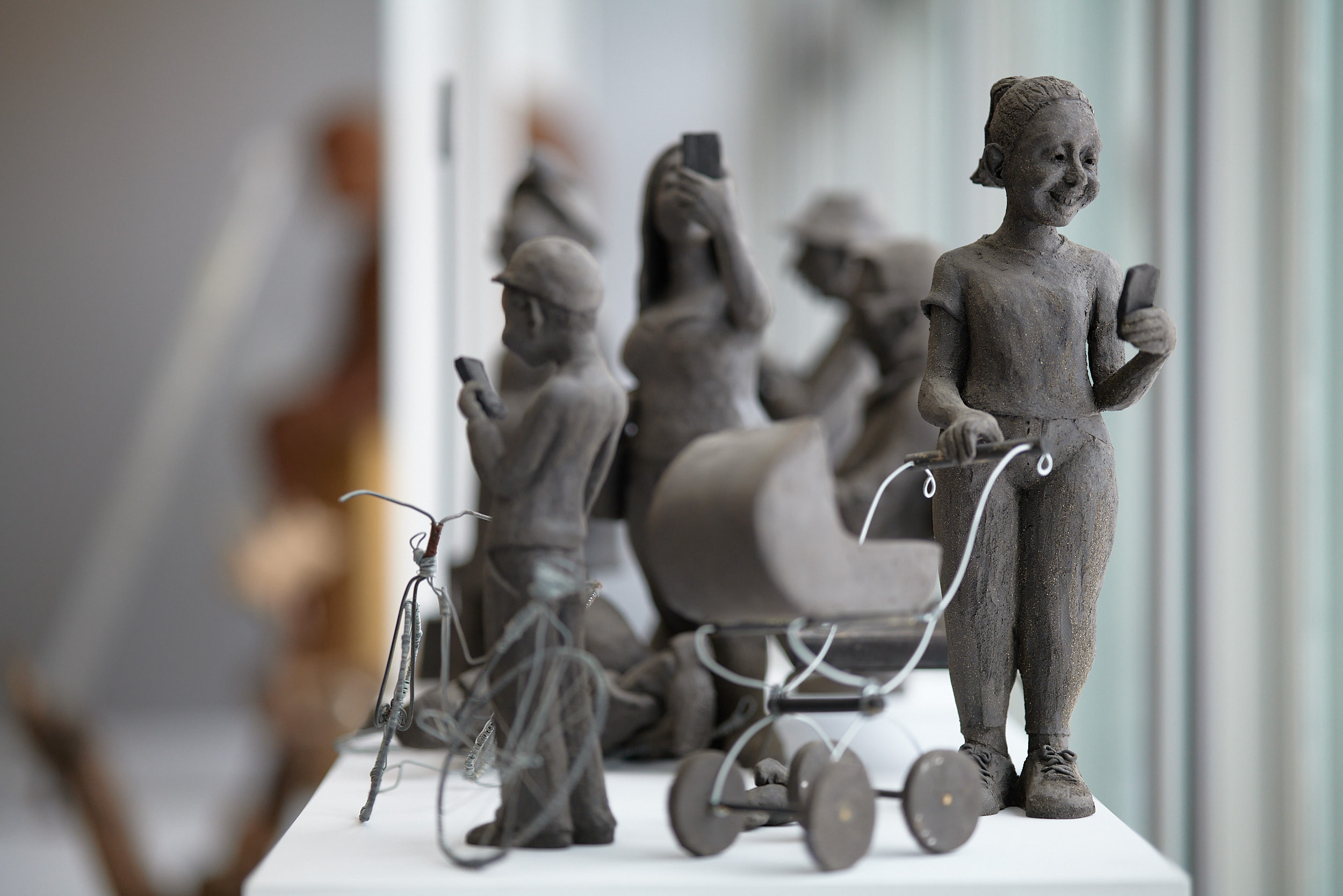 Närbild på en lerskulptur föreställande en större grupp människor. I förgrunden syns en barn vid en cykel och en kvinna vid en barnvagn. Båda tittar i sina mobiler.