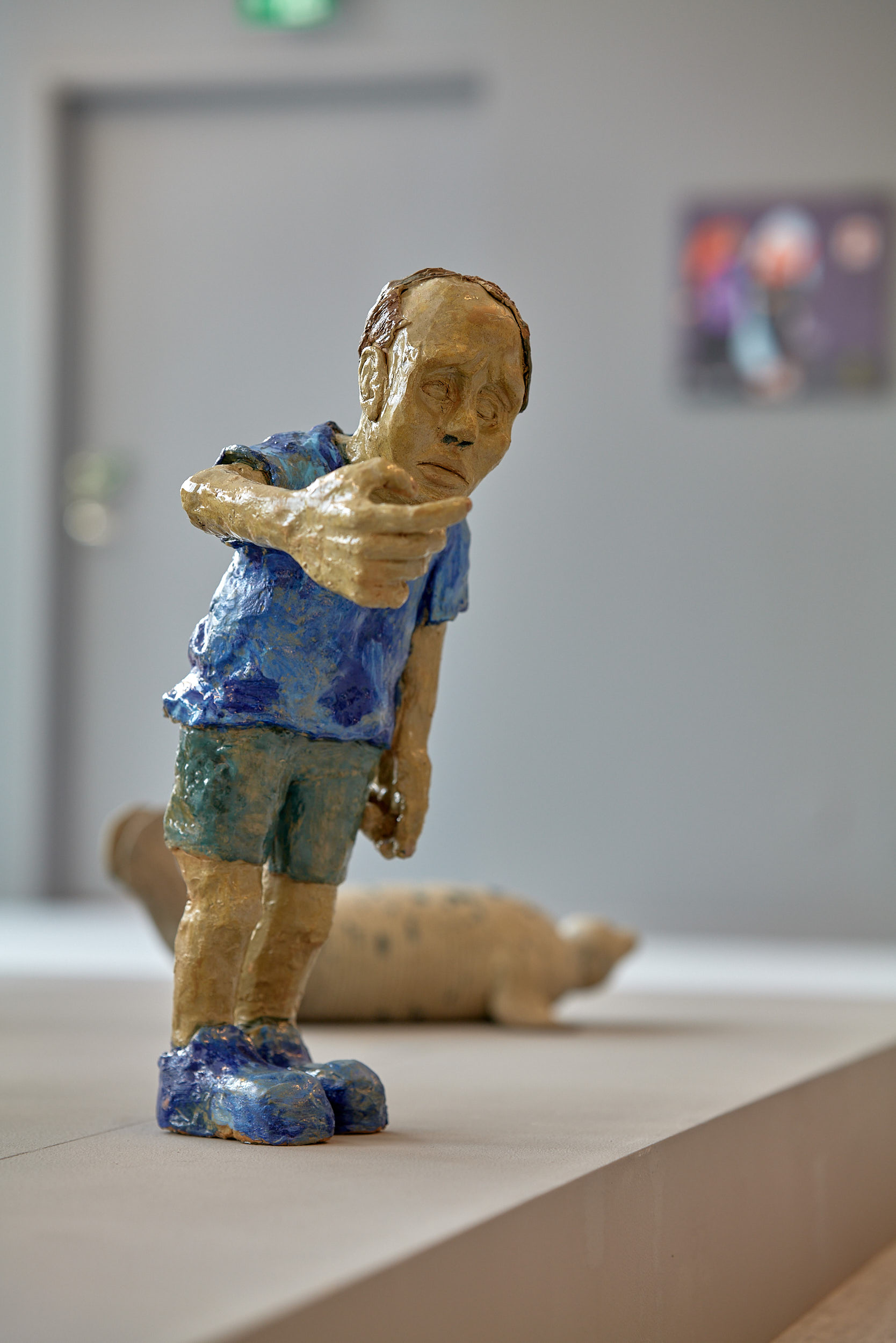 Fotografi på en keramikskultur föreställande en tunhårig man som står med höger hand lyft framför hakan. Mannen har blå skjorta, gröna shorts och blå skor.