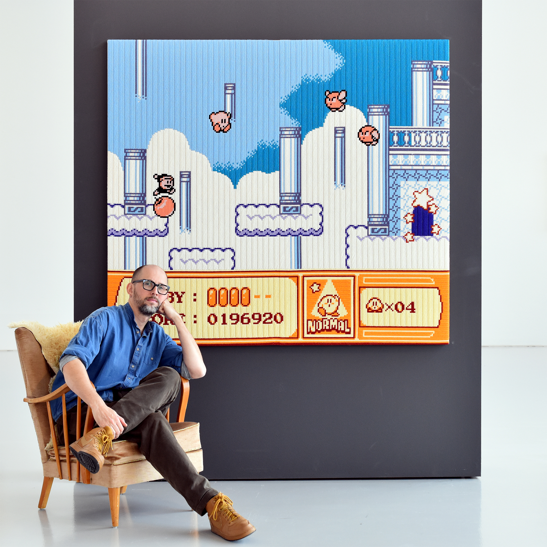 Fotografi på en man som sitter i en fotölj framför en fristående vägg. På väggen hänger en stor tavla med broderat motiv från ett retro-TV-spel. Mannen är skallig, har glaögon, mörkgrå byxor, blå skjorta och bruna skor.